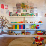 Organised play area with colorful storage shelves, educational materials, and tree wall decoration at Albany childcare
