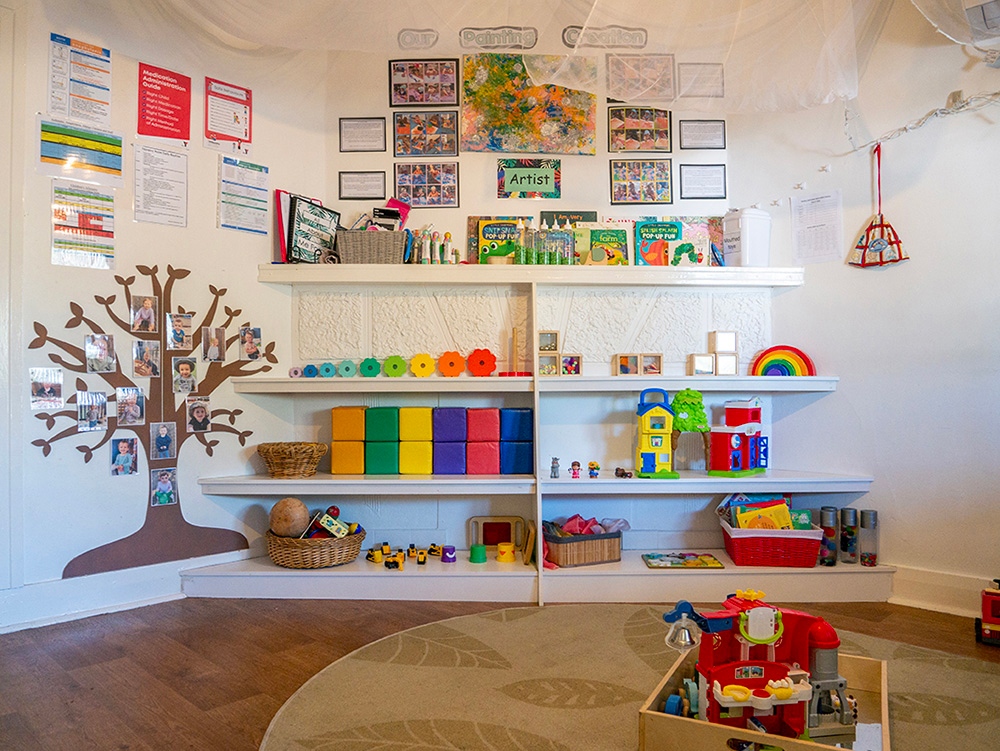 Organised play area with colorful storage shelves, educational materials, and tree wall decoration at Albany childcare
