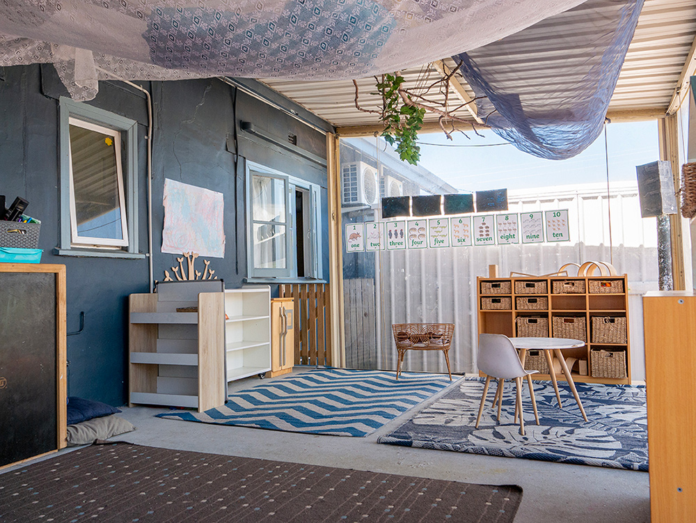 Covered outdoor learning space with wooden furniture, fabric canopy, and educational posters at Albany childcare centre