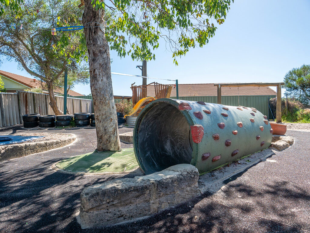 Large painted pipe climbing structure in outdoor play area at Albany childcare centre, with a tree and slide