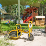Outdoor play area with yellow taxi trike, wooden fort with red slide, and shade sails at Baldivis childcare centre