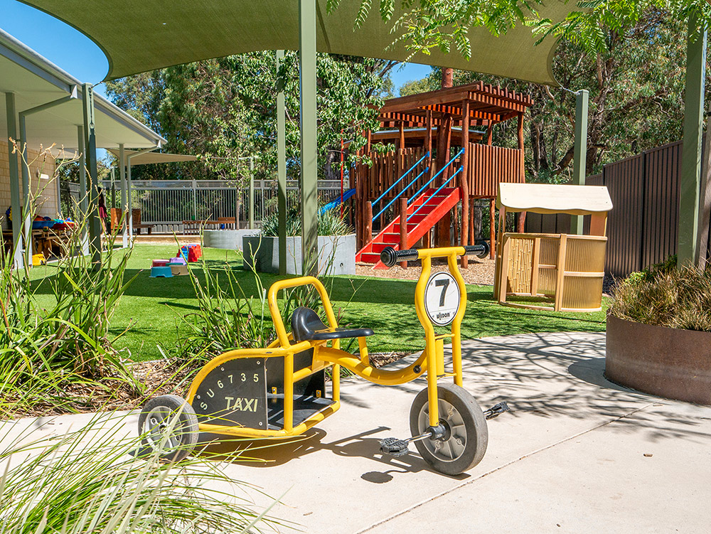 Outdoor play area with yellow taxi trike, wooden fort with red slide, and shade sails at Baldivis childcare centre