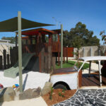 Outdoor play area with vintage car, wooden fort, sandpit, and shade sails at Brabham childcare centre