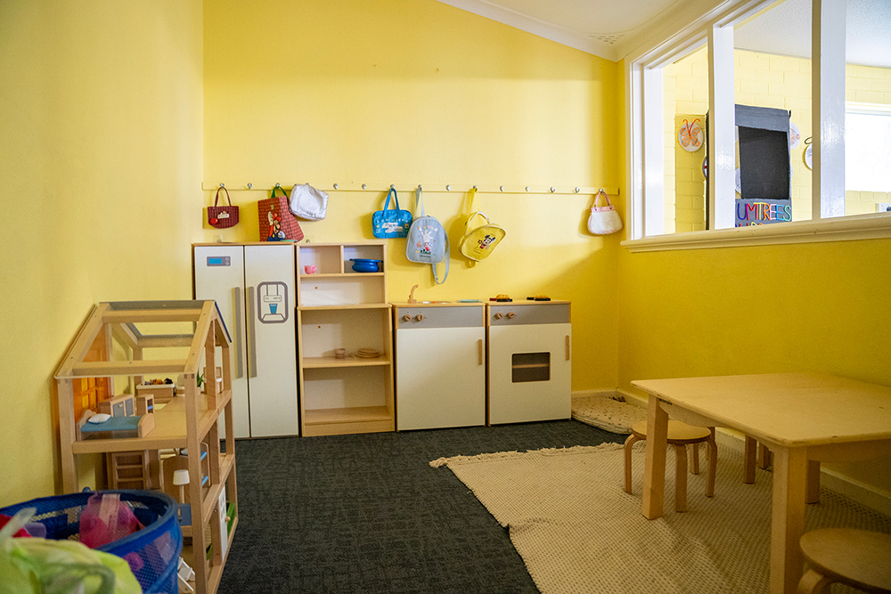 Play kitchen, dollhouse, and low tables in Gumtrees OSHC's yellow playroom with children's bags on wall hooks