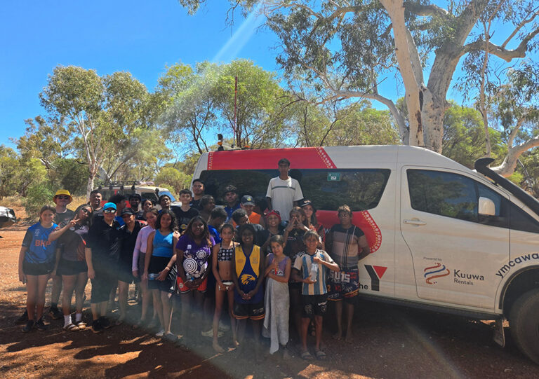 A large group of young people and adults pose together in front of a Y WA and Kuuwa Rentals van on a red dirt track surrounded by Australian bush