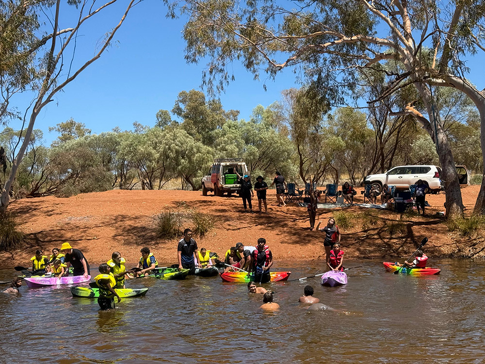 A group of young people kayaking and swimming in a river with red dirt banks and gum trees, with adults and vehicles visible on the bank behind them