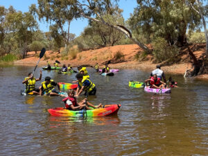 A group of children and young people in life jackets paddle colourful kayaks on a river surrounded by red dirt banks and gum trees on a sunny day