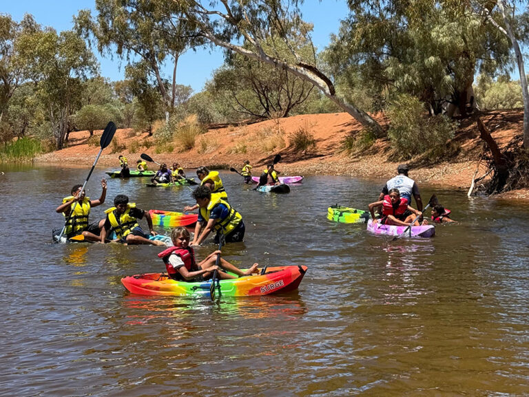 A group of children and young people in life jackets paddle colourful kayaks on a river surrounded by red dirt banks and gum trees on a sunny day