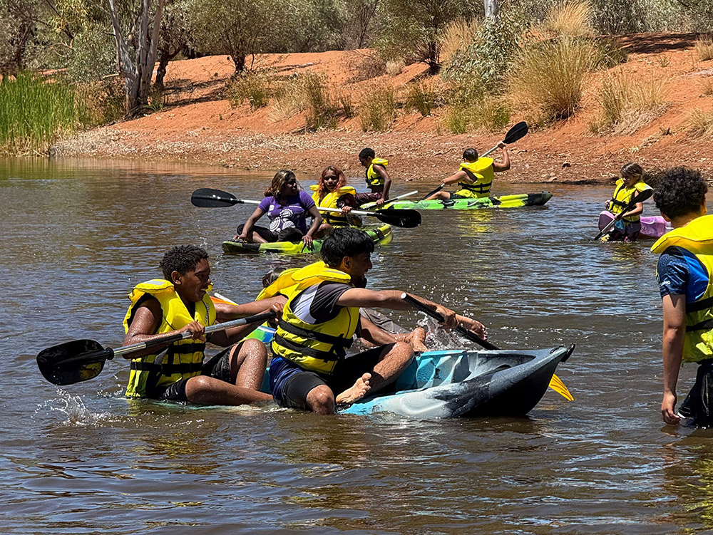 Young people in yellow life jackets paddle kayaks on a outback river with red sandy banks, with two in the foreground laughing as their kayak tips
