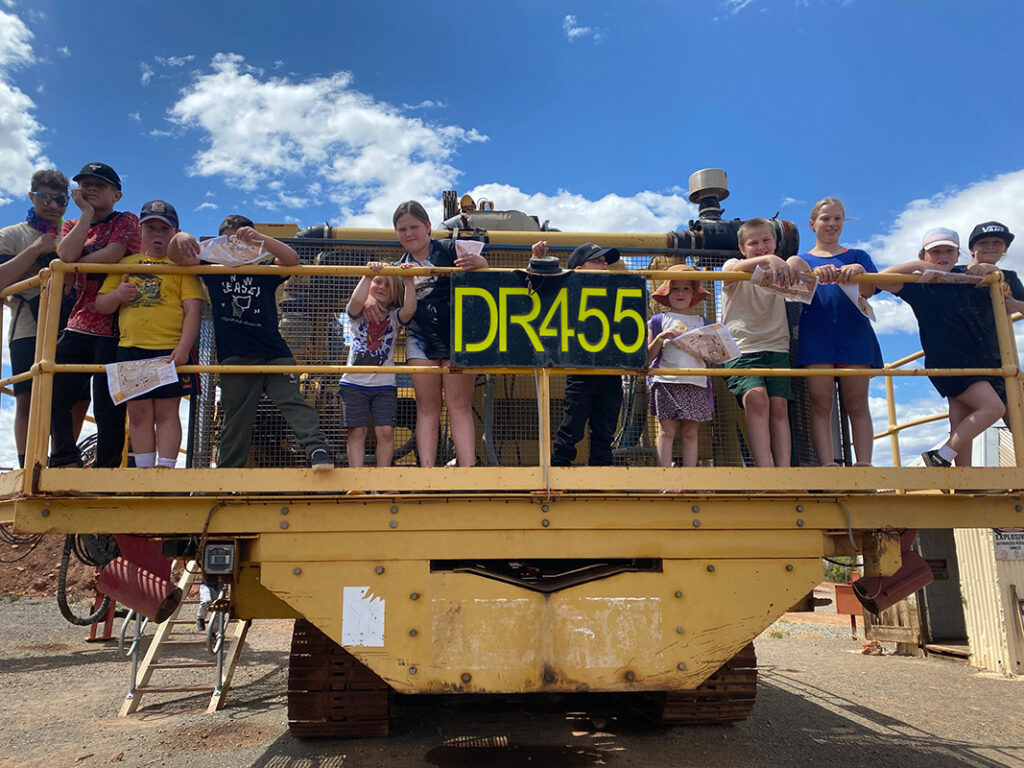 A group of children standing on a large yellow mining drill rig, labelled DR455, on a holiday program excursion under a blue sky