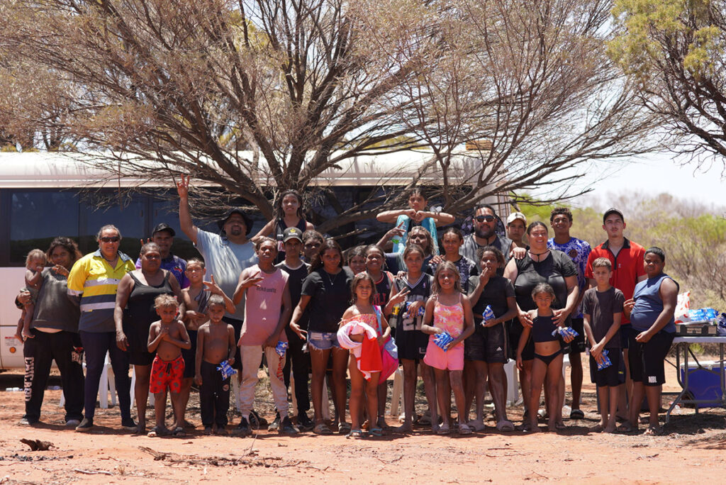 A large group of Aboriginal community members, adults and children, gathered outdoors near a bus and a tree on red dirt ground in a remote location