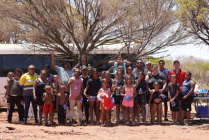 A large group of Aboriginal community members, adults and children, gathered outdoors near a bus and a tree on red dirt ground in a remote location