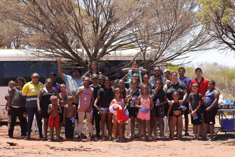 A large group of Aboriginal community members, adults and children, gathered outdoors near a bus and a tree on red dirt ground in a remote location