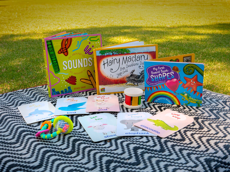 A collection of early childhood resources displayed on a patterned rug on grass, including children's books, animal sound flashcards, and small sensory toys