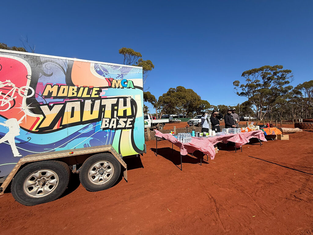 A colourfully wrapped mobile youth base trailer parked on red dirt, with trestle tables set up alongside it for a community event