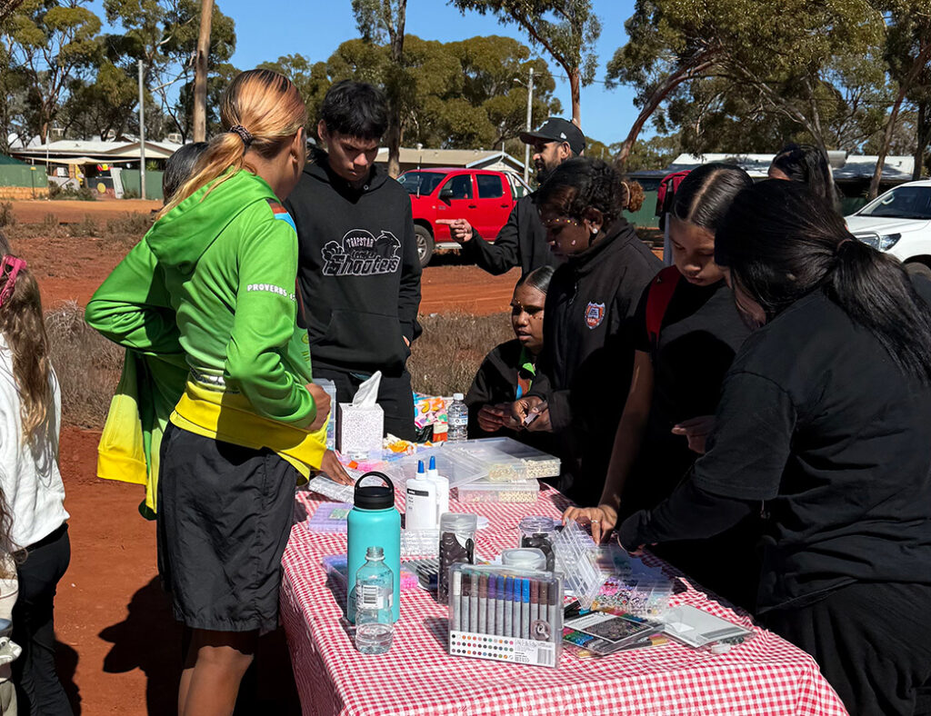 A group of young people gathered around a trestle table doing a craft activity outdoors on red dirt, supervised by a youth worker in a green jacket