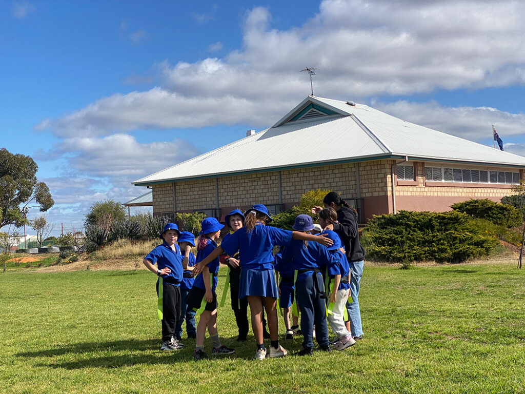 A group of children in blue school uniforms huddling together on a grassed oval with a school building in the background