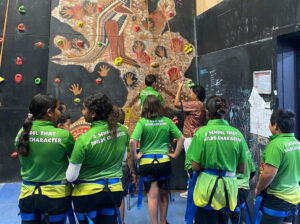 A group of children in green school uniforms watching a peer attempt a rock climbing wall decorated with Indigenous artwork