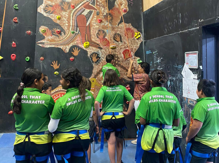 A group of children in green school uniforms watching a peer attempt a rock climbing wall decorated with Indigenous artwork