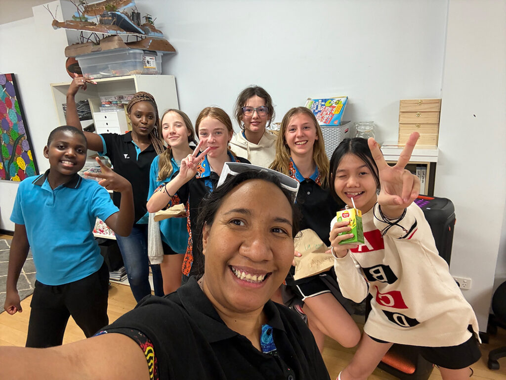 A youth worker takes a selfie with a smiling group of diverse young people giving peace signs during a Y WA program session indoors