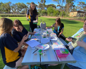 A group of young people and two youth workers doing watercolour painting at an outdoor picnic table on a grassed area