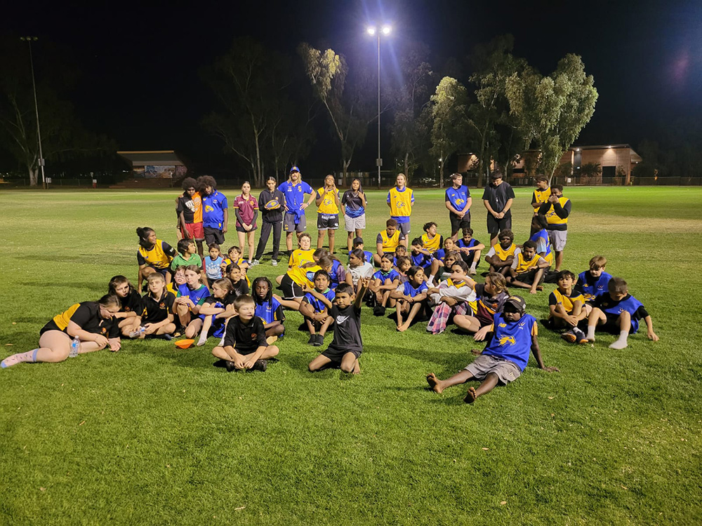 A large group of children and adults in West Coast Eagles bibs gather on a floodlit oval at night after an AFL session