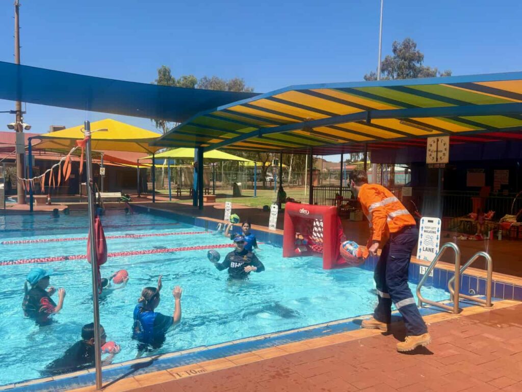 An adult in hi-vis workwear crouches poolside next to a group of children standing in an outdoor swimming pool, several holding AFL footballs
