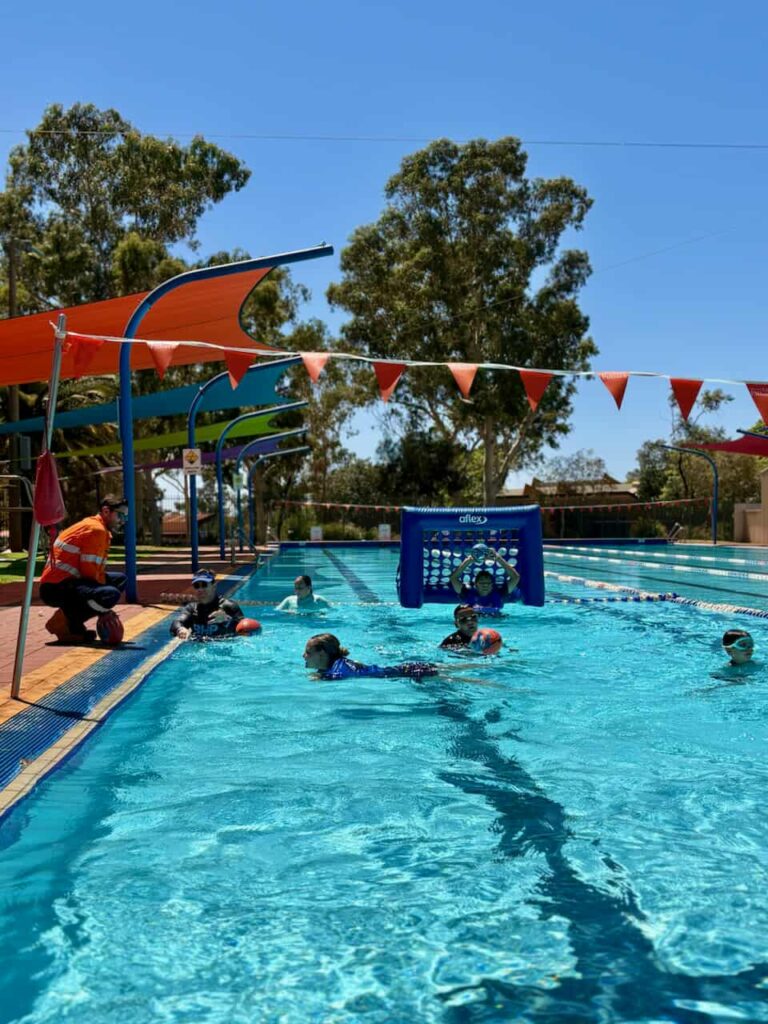 Children playing a water polo-style game in an outdoor pool, with an adult in hi-vis workwear supervising from the pool deck