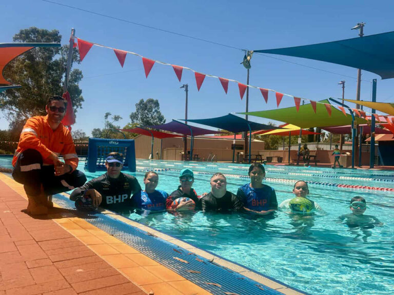Children swimming in an outdoor pool during a water sports activity session, with an inflatable goal in the water and an adult in hi-vis crouching at the pool's edge