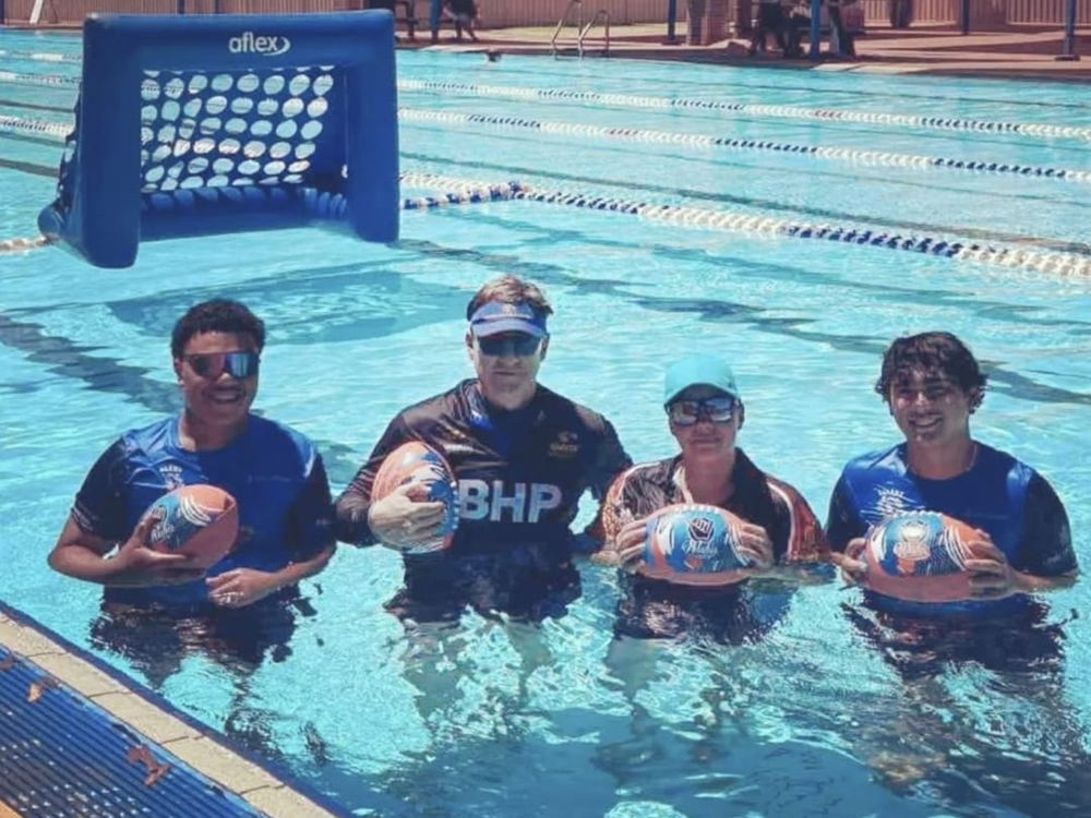 Four people standing in an outdoor swimming pool holding AFL footballs, with an inflatable goal in the background