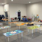 A bright, open children's care room set up with activity tables laid with colourful plates and learning materials, surrounded by play areas and bookshelves