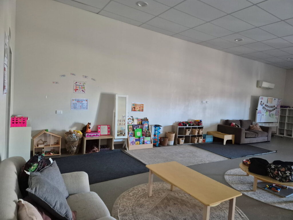 A spacious children's care room with sofas, a dollhouse, book display, toy shelves, and a pretend salon play area along the walls