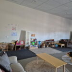 A spacious children's care room with sofas, a dollhouse, book display, toy shelves, and a pretend salon play area along the walls