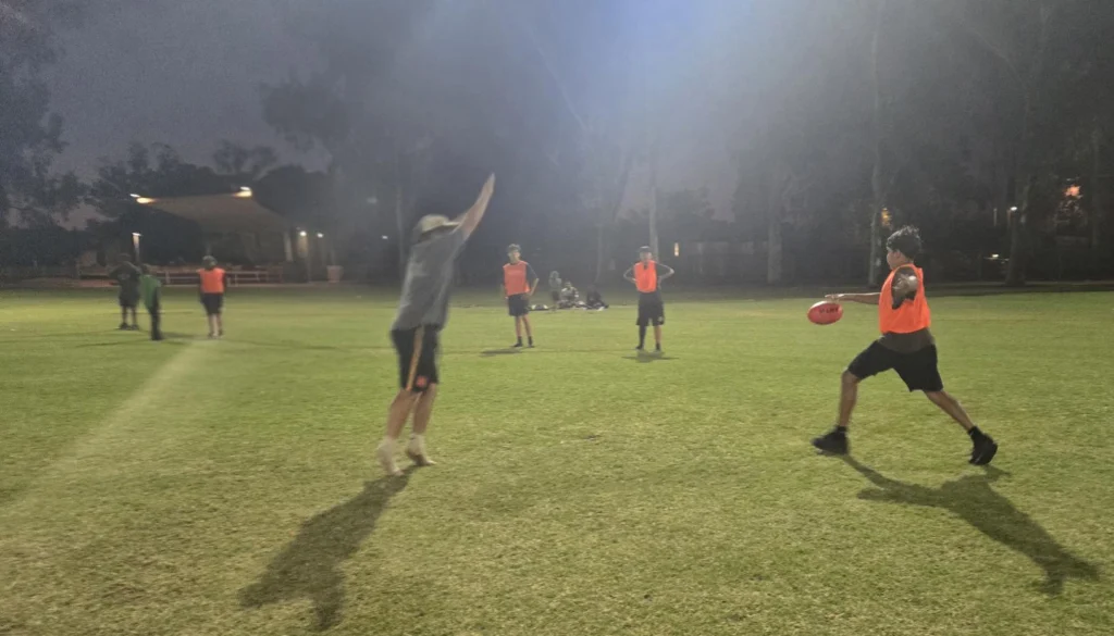Players in orange bibs practise kicking and marking a football under floodlights on a dark oval at night