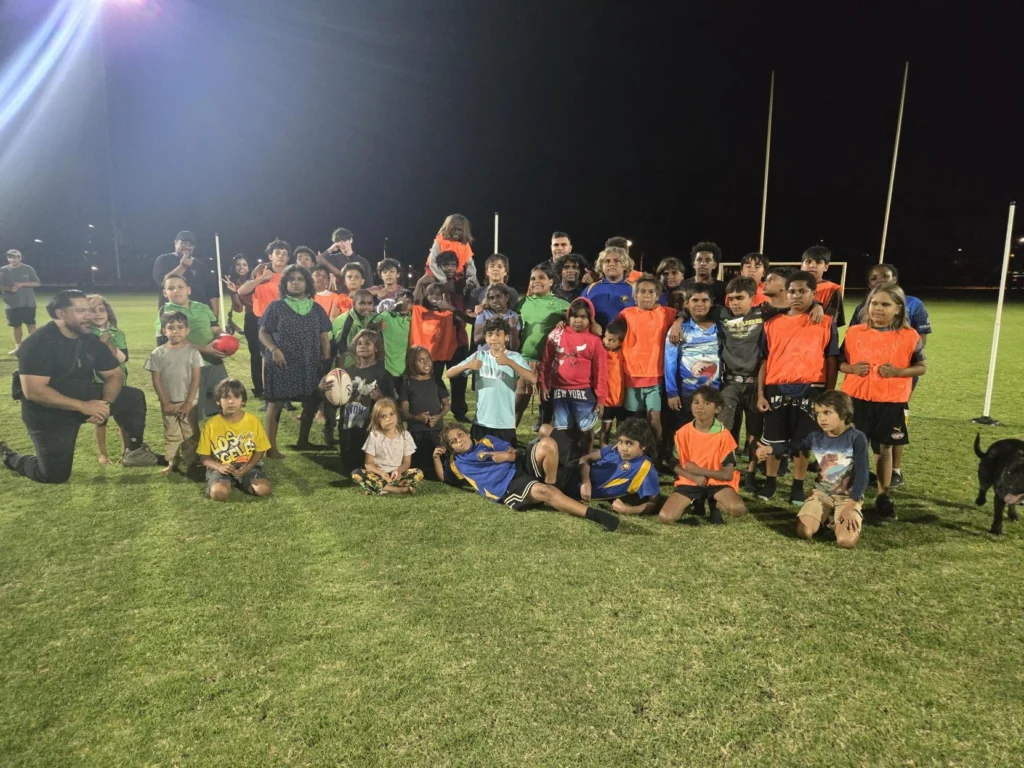A large group of children and adult coaches pose together for a team photo on a football oval at night, many wearing orange and green training bibs, with goal posts and a dog in the background