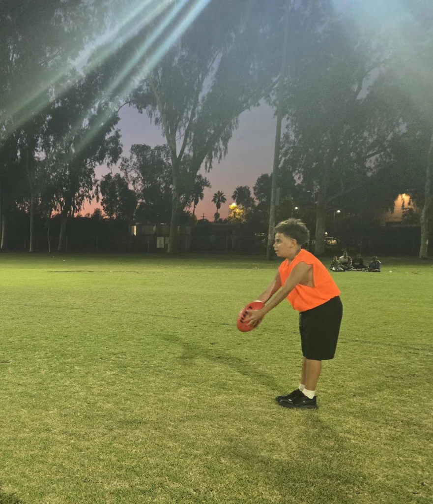 A young player in an orange bib prepares to kick a football on a grass oval at dusk, with trees and spectators seated in the distance