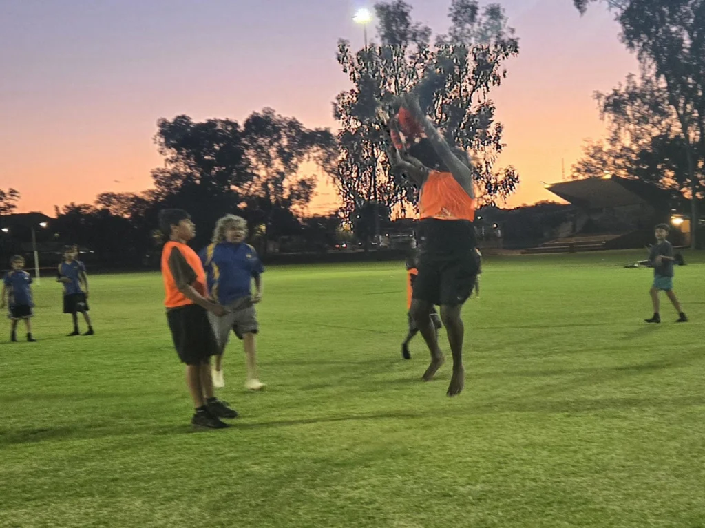 A player in an orange bib leaps to catch a football mid-air on a sports field at sunset, with teammates and a grandstand visible in the background