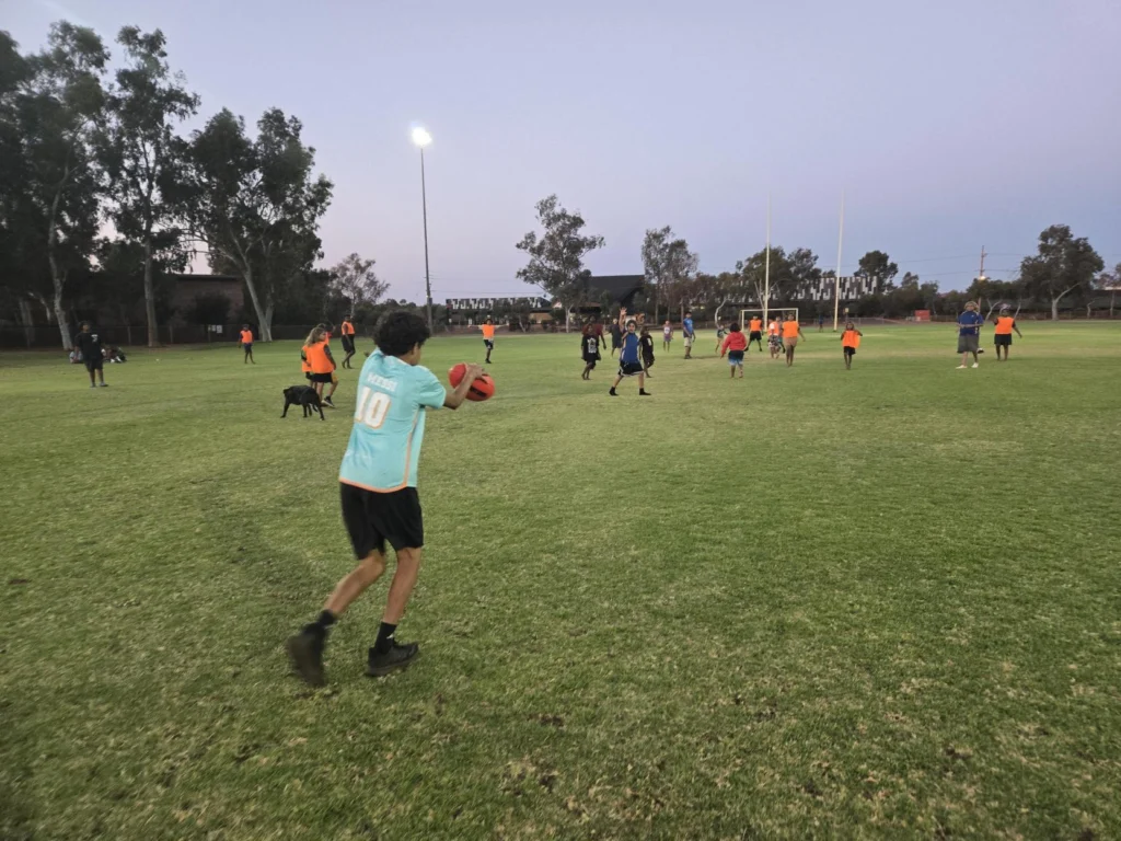 A player in a teal jersey holds an Australian rules football, running across a grass oval at dusk with other players and a dog scattered across the field, floodlight and goal posts visible in the background.