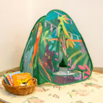 A jungle-themed pop-up play tent with a wicker basket of children's books beside it, set on a patterned rug in an early learning centre
