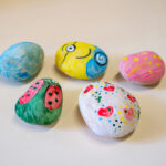 Five colorful painted rocks created by children featuring emoji, ladybug and heart designs