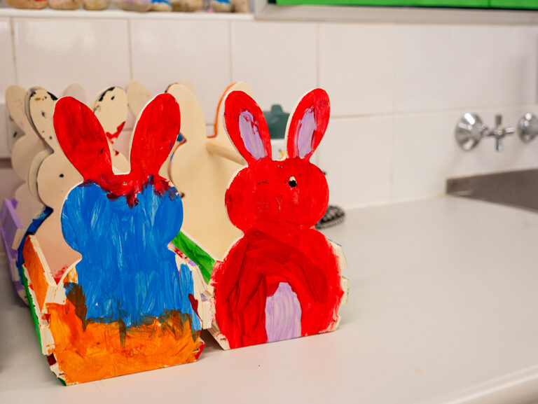 Two hand-painted wooden Easter bunny shapes in red and blue sitting on a bench near a sink