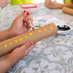 A child's hands paint yellow and red dots onto a cardboard tube in an Indigenous-inspired craft activity