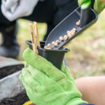 A gloved child holds a small pot as seeds are poured in from a trowel during a planting activity.