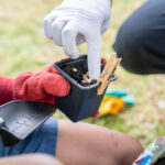 A child's gloved hands hold a small pot while an adult in gloves places seeds into the soil