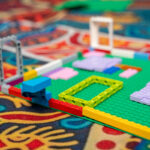 A LEGO build in progress on a green baseplate, featuring colourful bricks arranged on a patterned classroom carpet