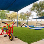 A red children's tricycle in the foreground of a shaded outdoor play area with climbing equipment and crash mats on artificial grass