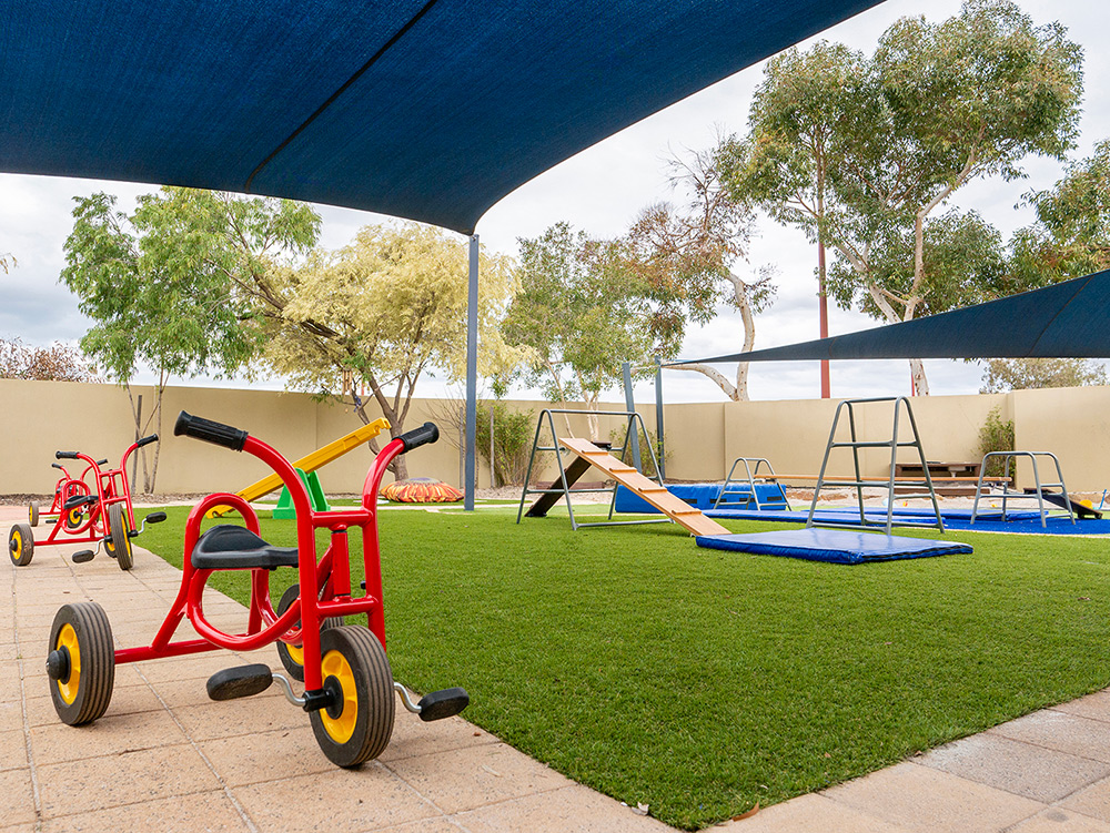 A red children's tricycle in the foreground of a shaded outdoor play area with climbing equipment and crash mats on artificial grass