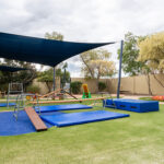 An outdoor play area with shade sails, balance beams, climbing frames, blue crash mats, and ride-on tricycles on artificial grass