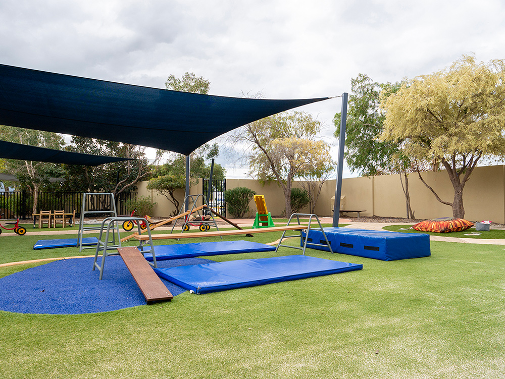 An outdoor play area with shade sails, balance beams, climbing frames, blue crash mats, and ride-on tricycles on artificial grass