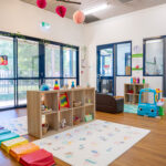 A light-filled early learning room with an alphabet play mat, wooden toy shelves, rainbow floor cushions, and large windows overlooking a garden